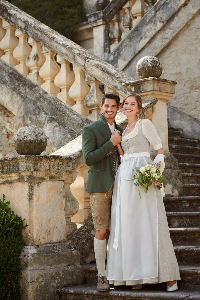 Trachtenhochzeit im Hochzeitsdirndl in hellem Beigeton und Bräutigam in elegantem Leinensakko und Hirschlederhose.