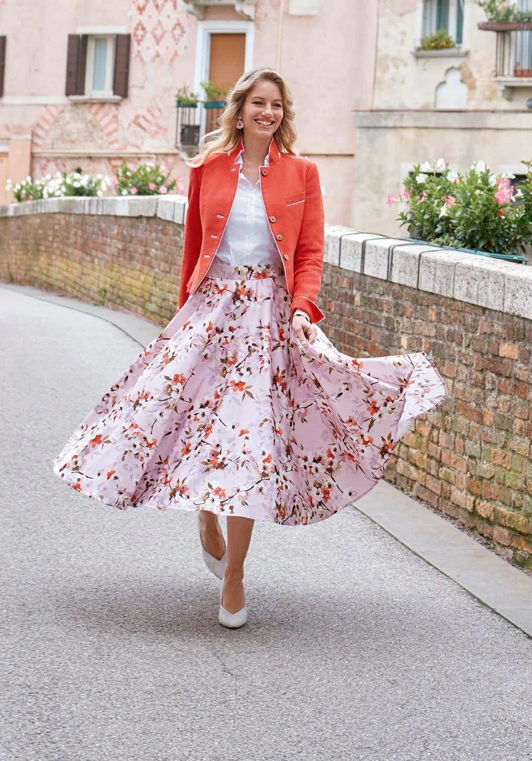 Blonde Frau in apricotfarbener Jacke und floralem Maxirock mit weißen Pumps in italienischer Gasse – Wimmer schneidert