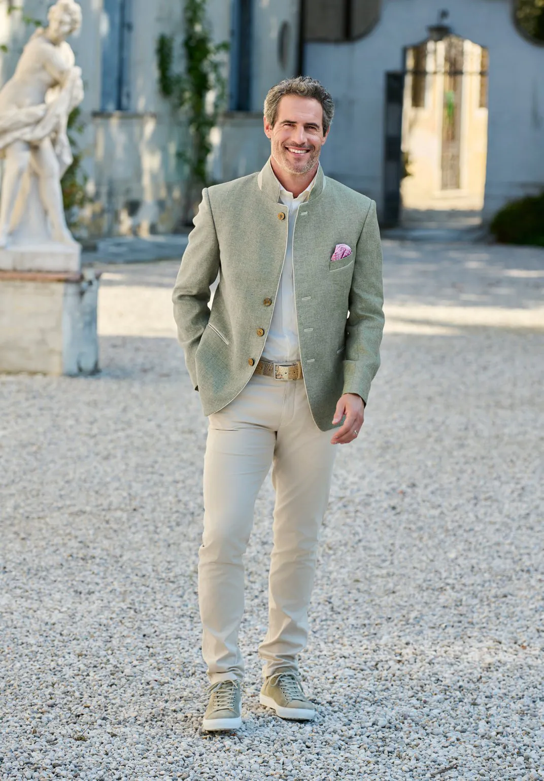 Herr in salbeifarbenem Leinen-Trachtenjanker mit rosa Einstecktuch und beiger Hose im Villeninnenhof – Wimmer schneidert