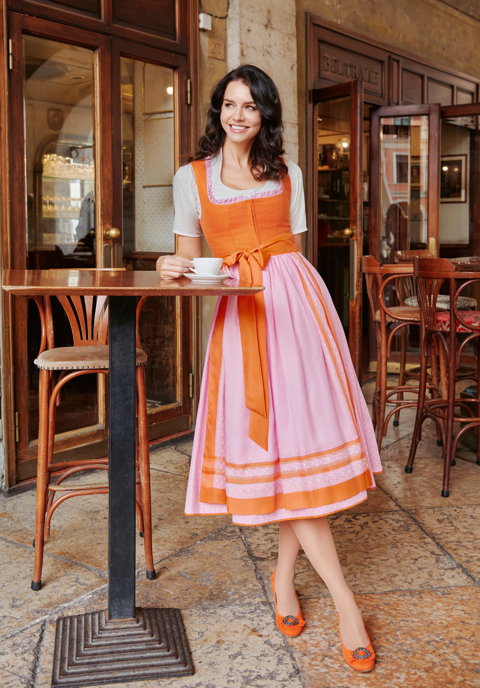 Frau in orangem Dirndl mit rosa Schürze an einem italienischen Café-Tisch mit Kaffeetasse – Lebendige Farben von Wimmer schneidert