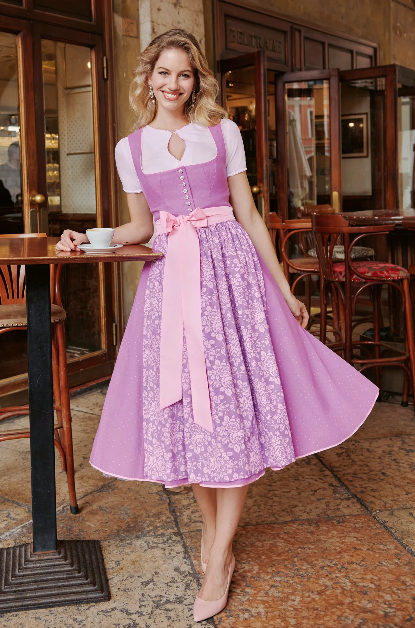 Blonde Frau in fliederfarbenem Dirndl mit floraler Lavendelschürze und rosa Satinschleife im Café – Wimmer schneidert