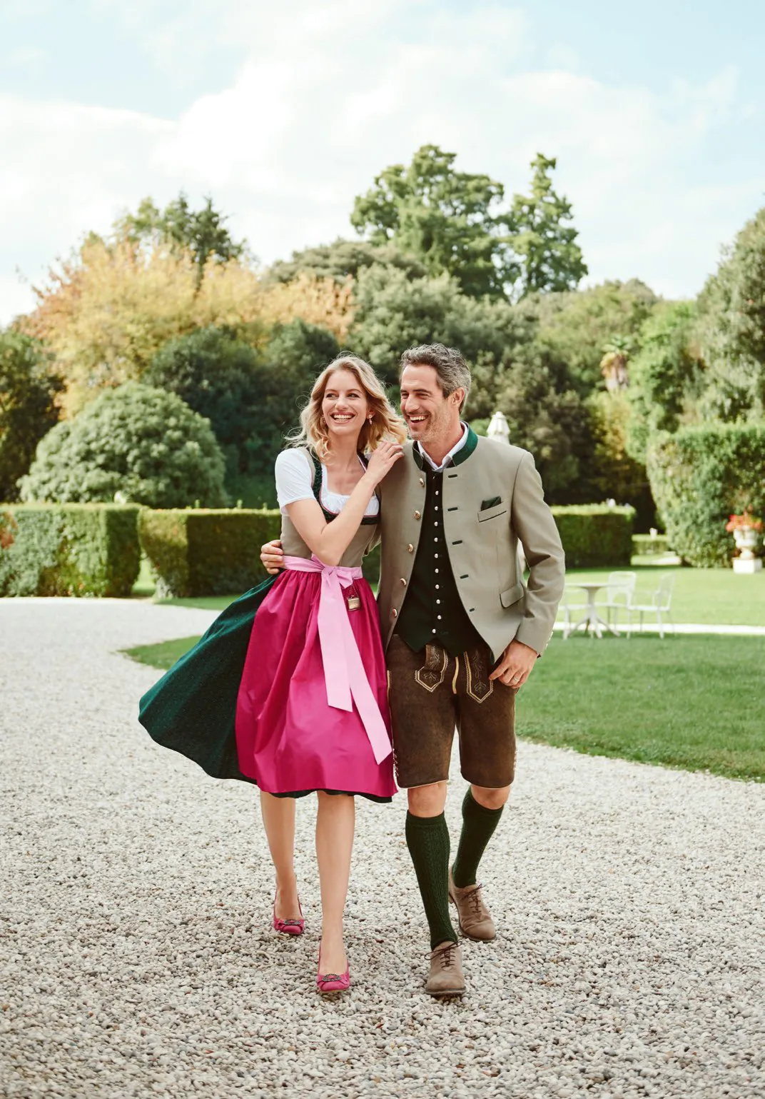 Paar spaziert im Garten – sie in fuchsiafarbenem Dirndl, er in Trachtenjanker und Lederhose – Tradition von Wimmer schneidert