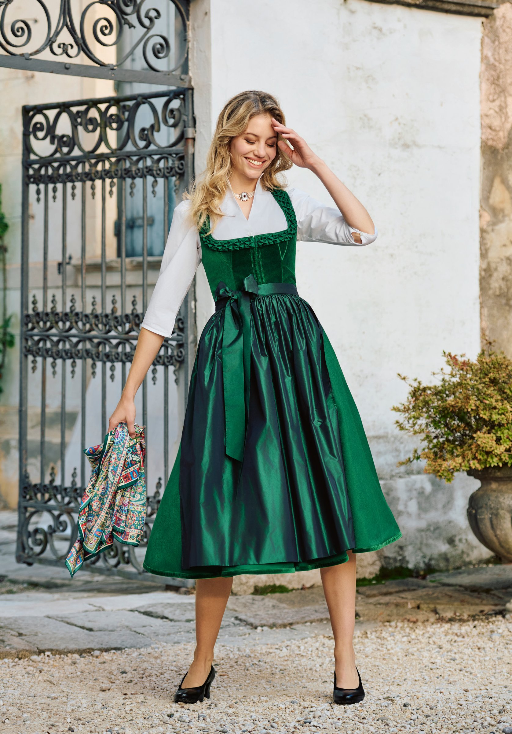 Blonde Frau in smaragdgrünem Samtdirndl mit grüner Satinschürze und buntem Seidenschal vor Villentor – Wimmer schneidert