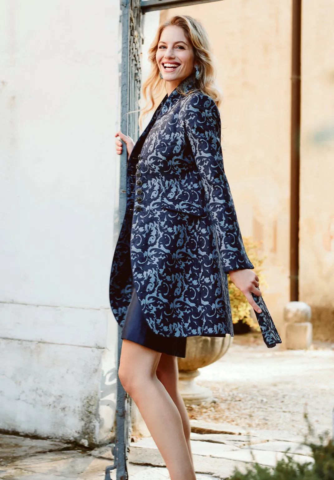 Elegante Frau in barockem Marinemantel und dunklem Kleid an einem goldbeleuchteten Türrahmen – Wimmer schneidert Lookbook