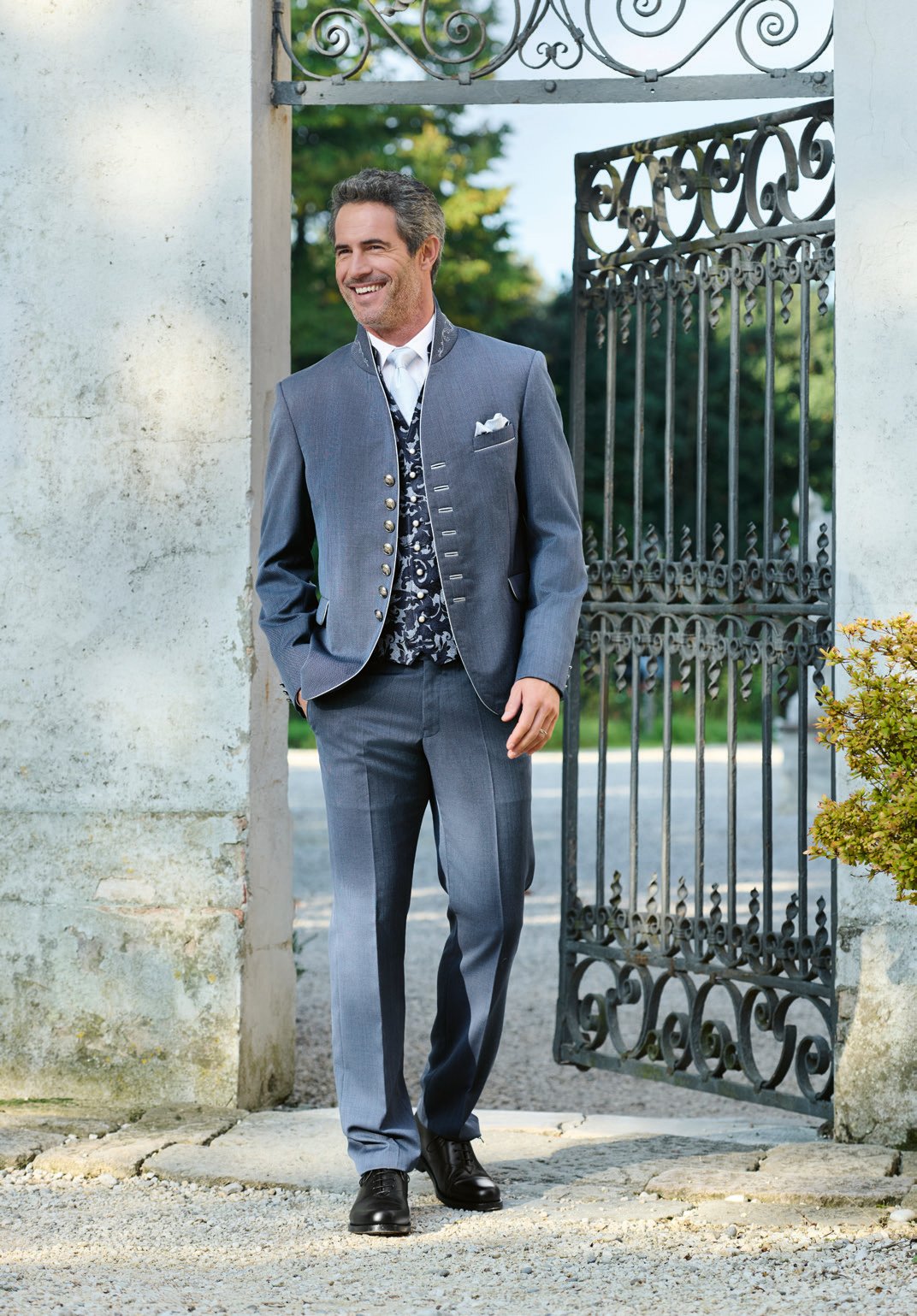 Herr in stahlblauem Trachtenanzug mit Brokatweste vor schmiedeeisernem Tor – Klassisch elegant von Wimmer schneidert