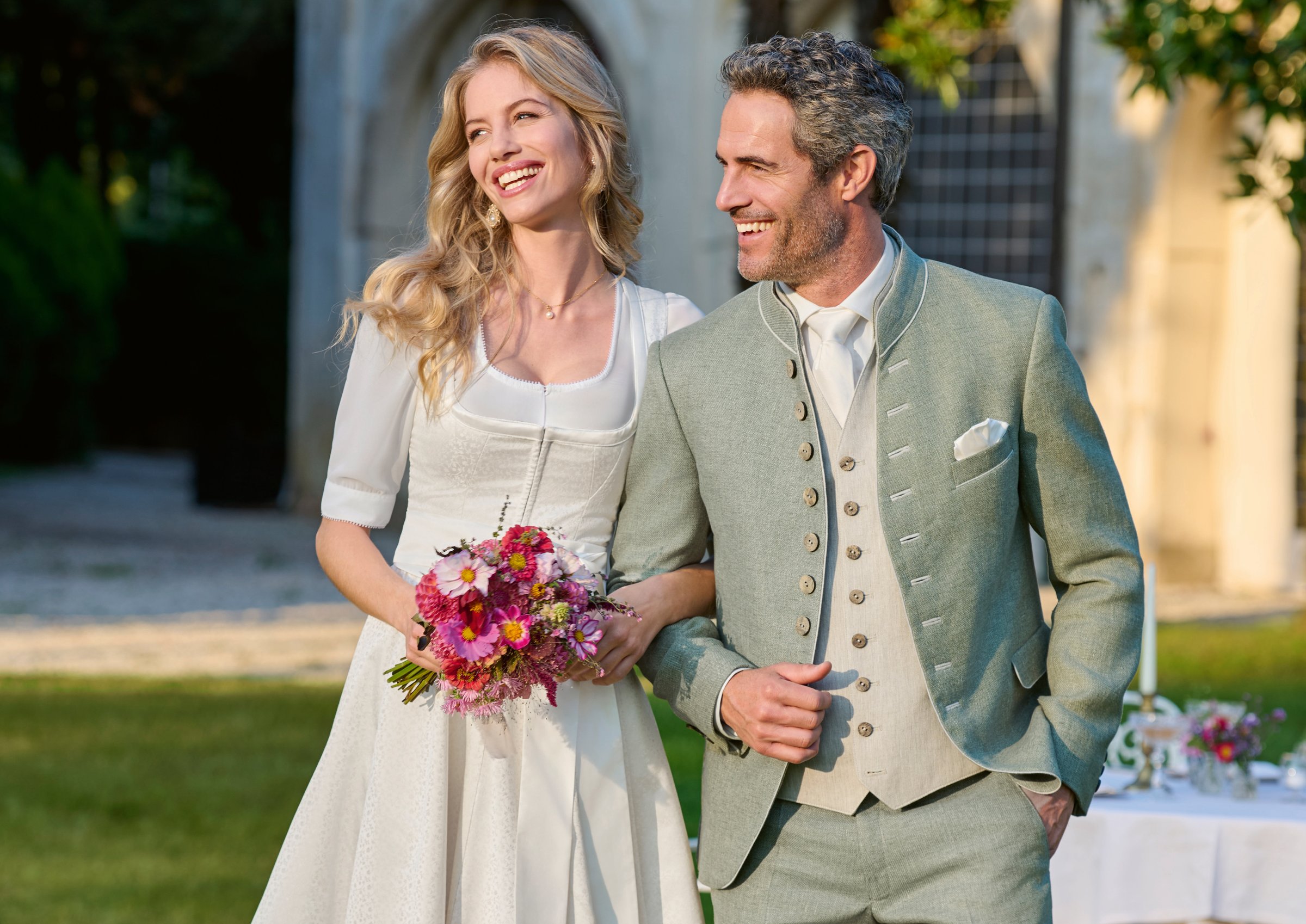 Strahlendes Hochzeitspaar vor Villenkapelle – Braut in weißem Hochzeitsdirndl mit Blumenstrauß, Bräutigam in salbeigrünem Trachtenanzug – Wimmer schneidert