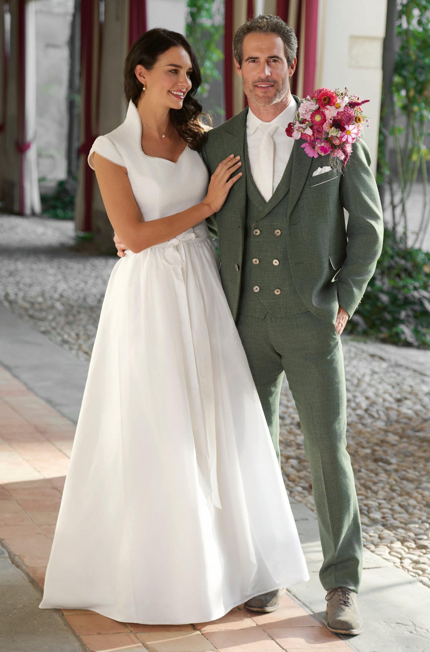 Hochzeitspaar – Braut in elegantem weißem Kleid mit breitem Kragen, Bräutigam in olivgrünem Dreiteiler mit rosa Strauß – Wimmer schneidert