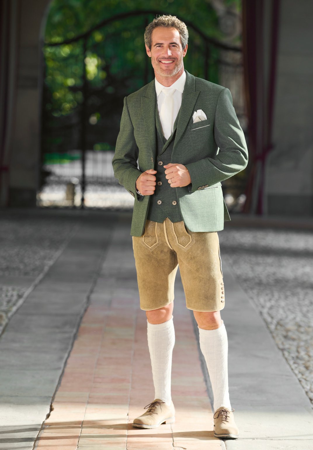 Bräutigam in olivgrünem Leinenanzug mit heller Lederhose und weißen Kniestrümpfen im Innenhof – Wimmer schneidert Hochzeit