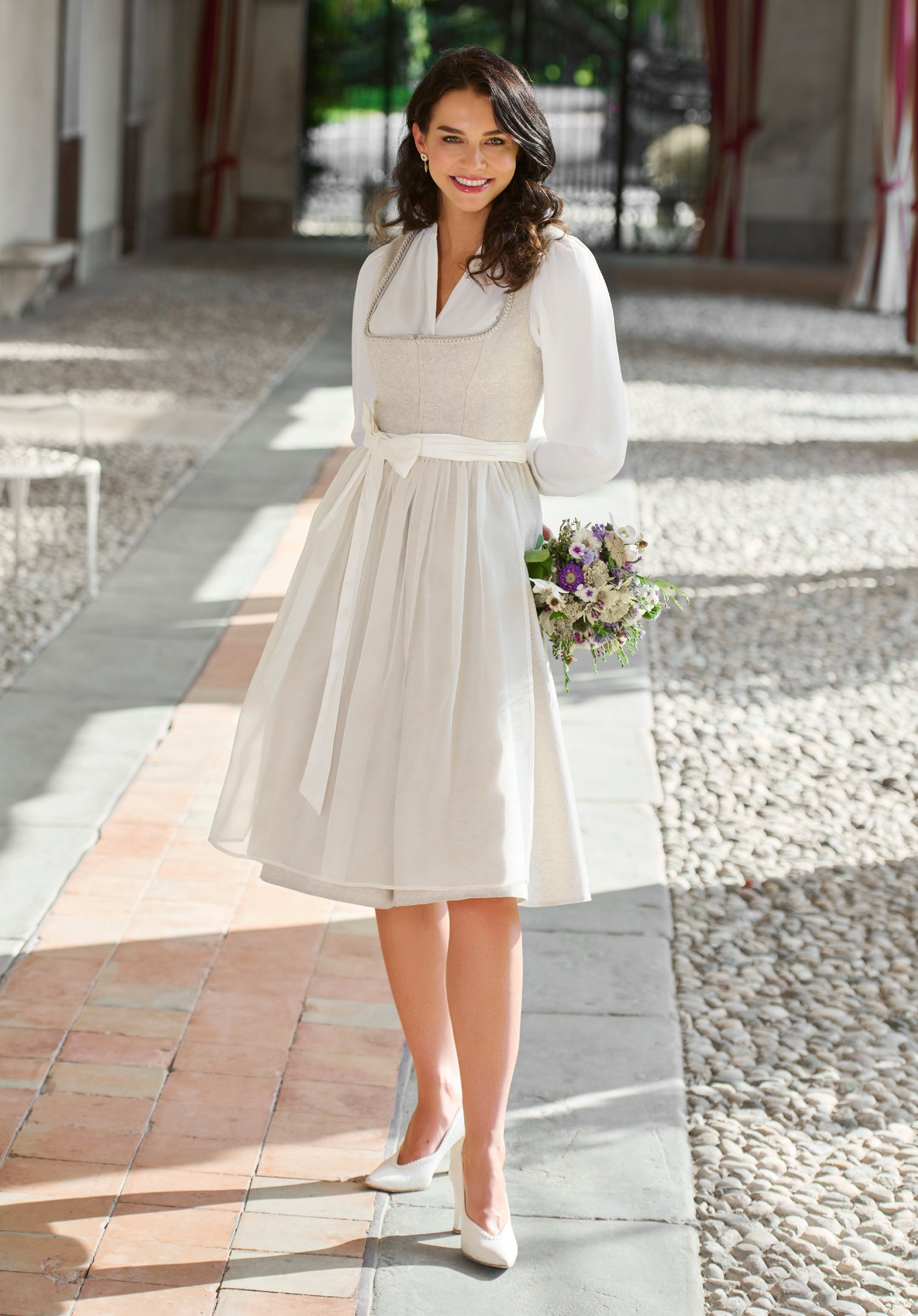 Braut in cremefarbenem Hochzeitsdirndl mit langen Chiffonärmeln und Lavendelstrauß im italienischen Innenhof – Wimmer schneidert