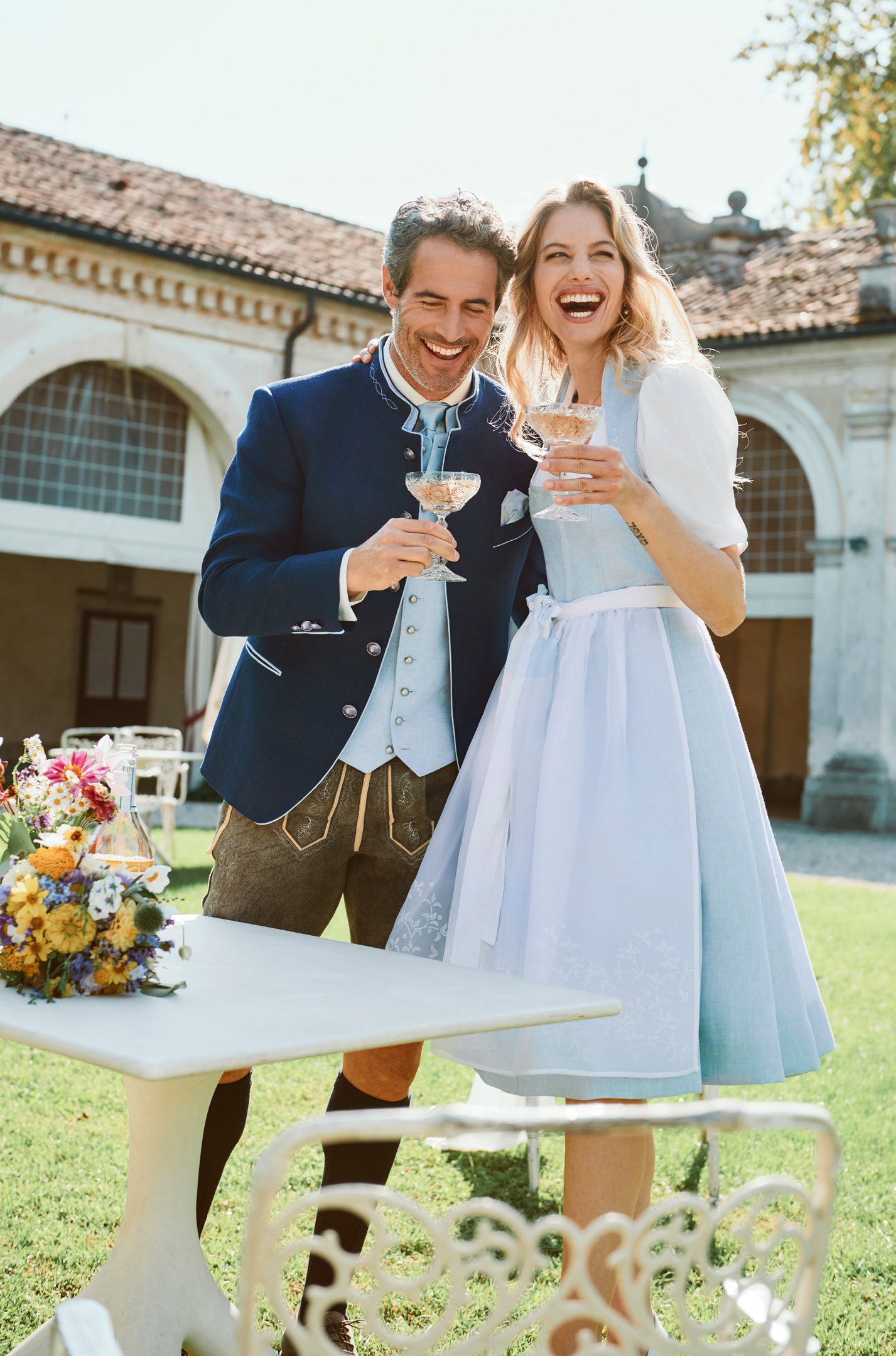 Paar stößt mit Champagner an – sie in hellblauem Dirndl mit weißer Schürze, er in Navy-Janker mit hellblauer Weste – Wimmer schneidert