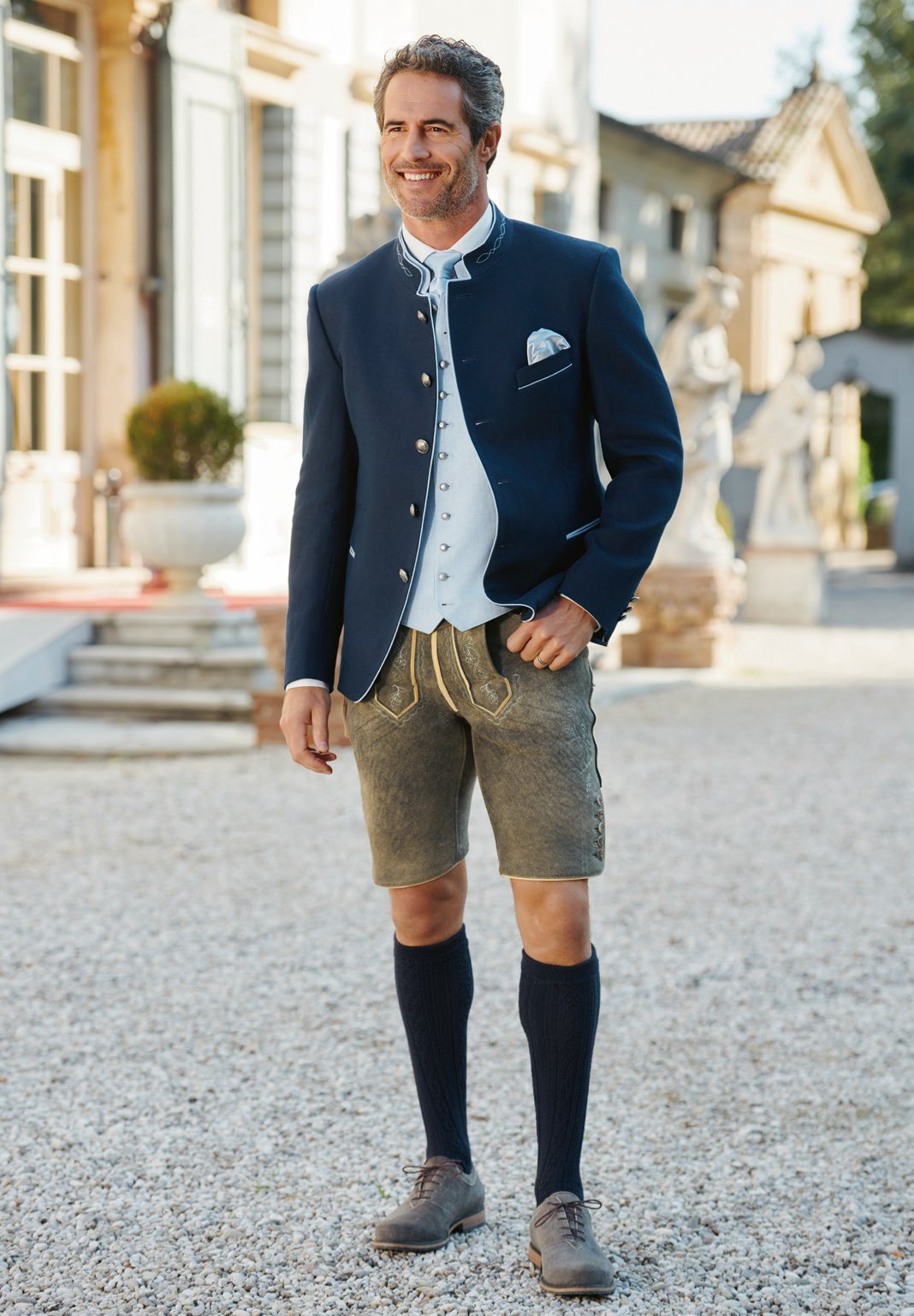 Herr in Navy-Trachtenjanker mit hellblauer Weste und olivfarbener Lederhose im Villeninnenhof – Wimmer schneidert