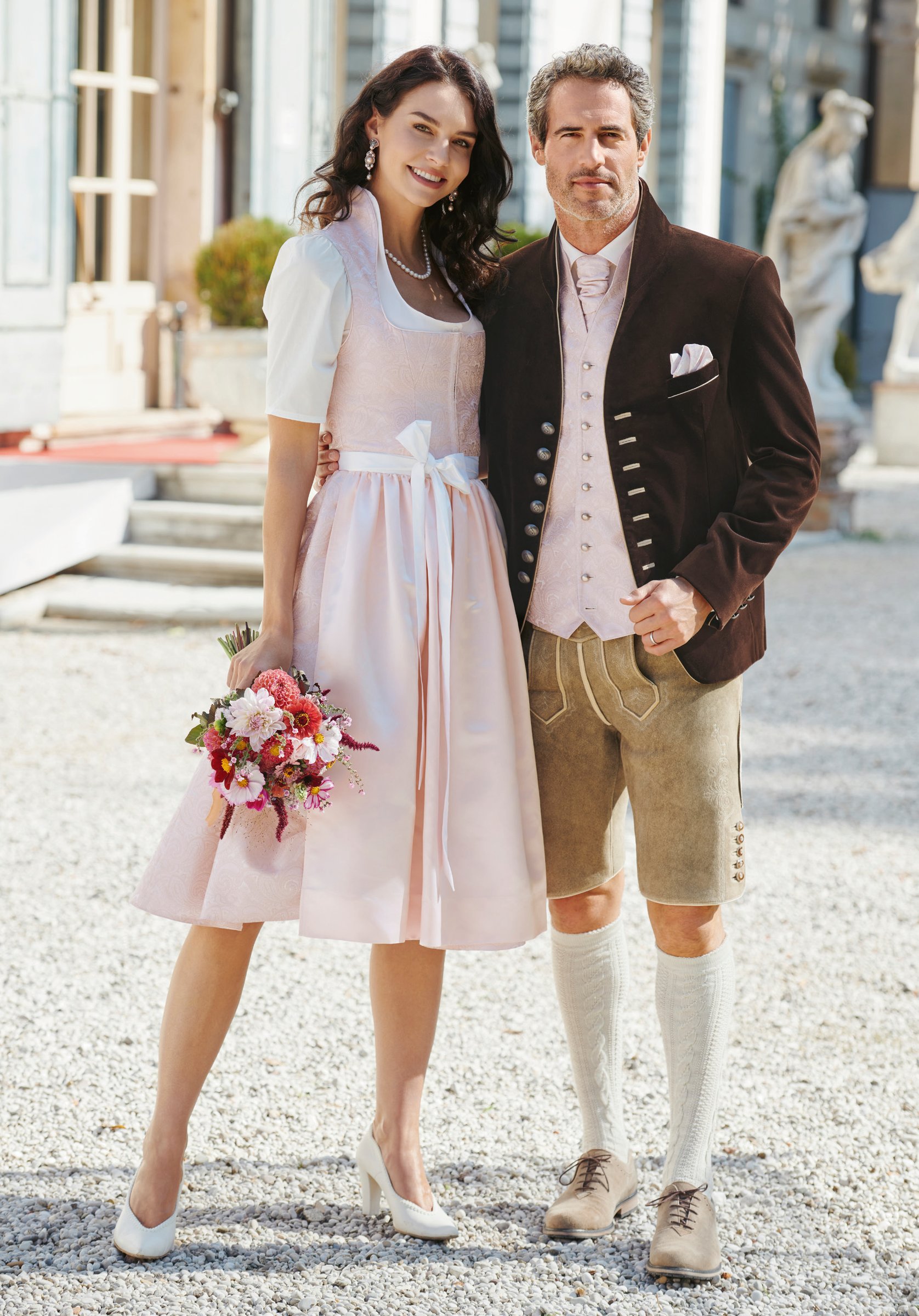 Paar im Villeninnenhof – sie in zartrosa Dirndl mit Blumenstrauß, er in burgunderrotem Samtjanker und Lederhose – Wimmer schneidert