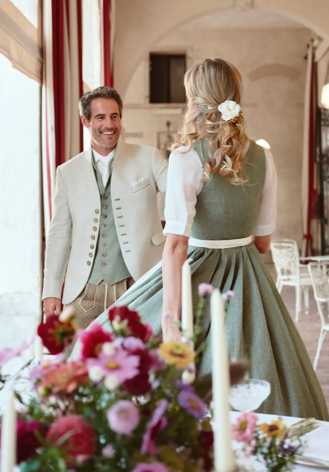 Paar von hinten an festlicher Tafel – sie zeigt salbeigrünes Dirndl mit Blumenhaarschmuck, er in cremefarbenem Janker – Wimmer schneidert