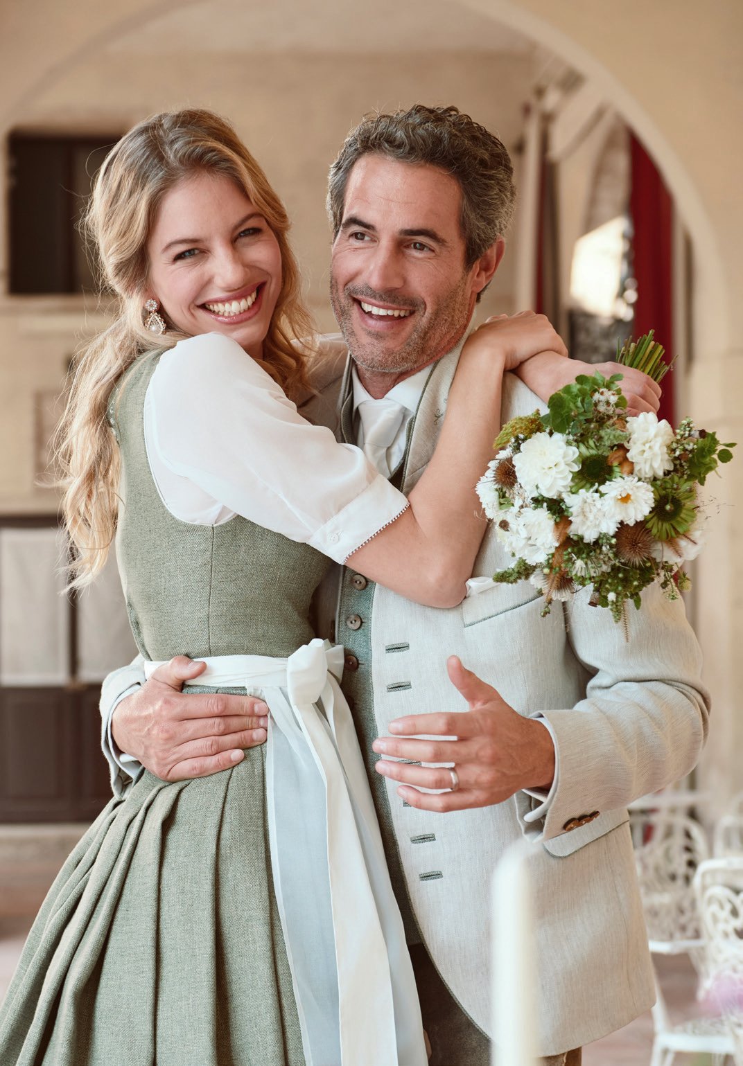 Verliebtes Paar umarmt sich – sie in salbeigrünem Dirndl, er in cremefarbenem Leinenanzug, mit Blumenstrauß – Wimmer schneidert