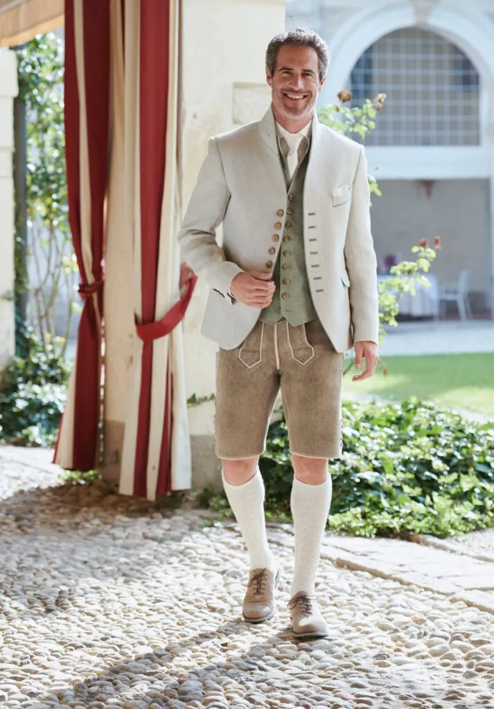 Herr in cremefarbenem Trachtenjanker, salbeigrüner Weste und goldener Lederhose im Villengarten