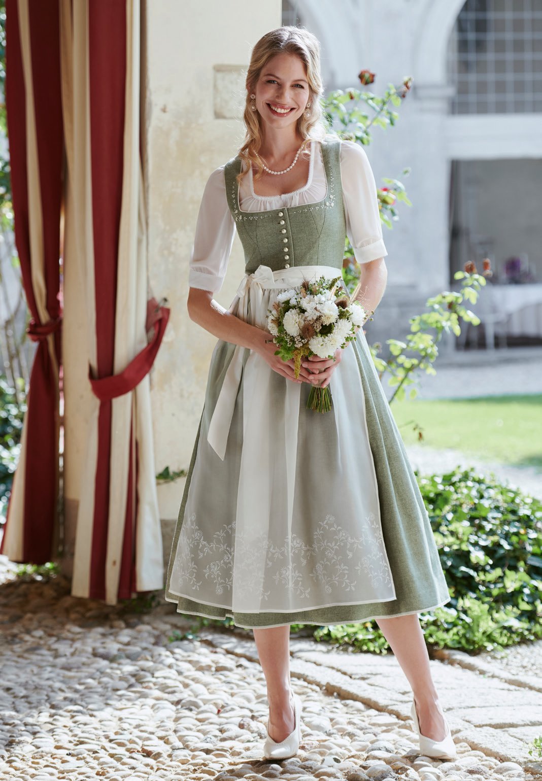Blonde Frau in salbeigrünem Brokatdirndl mit cremefarbener Edelweiß-Schürze und weißem Blumenstrauß im Villengarten – Wimmer schneidert