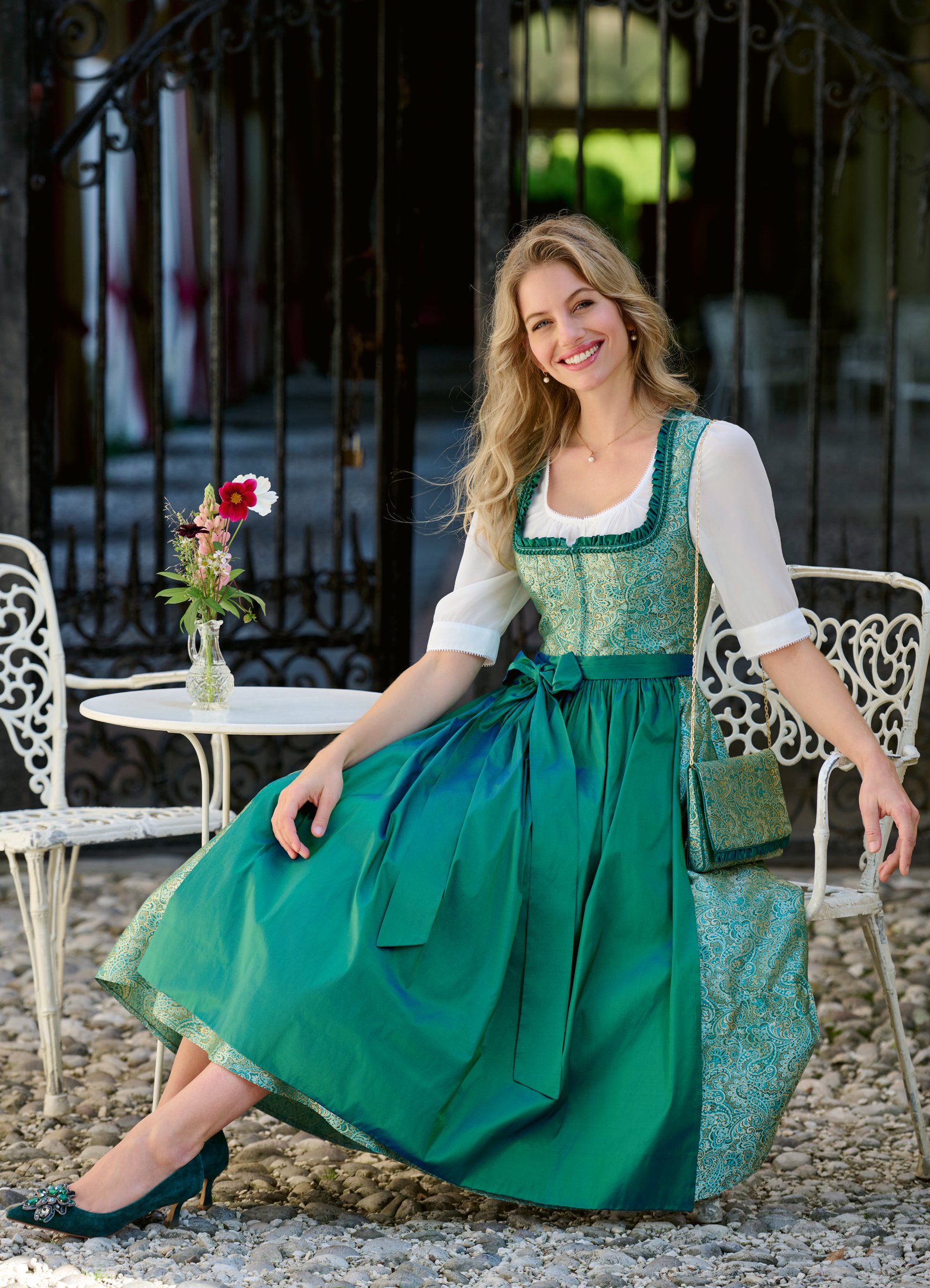Lächelnde Frau in smaragdgrünem Brokatdirndl mit grüner Satinschürze an weißem Gartencafé-Tisch mit Blumenvase – Wimmer schneidert