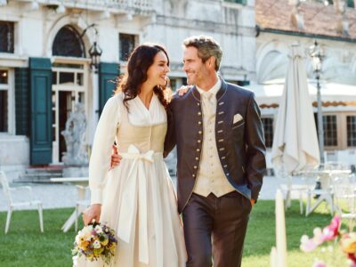 Traditionsbrautpaar spaziert durch Schlossgarten - elegant in Trachtenkleidung mit historischer Schlossarchitektur im Hintergrund.