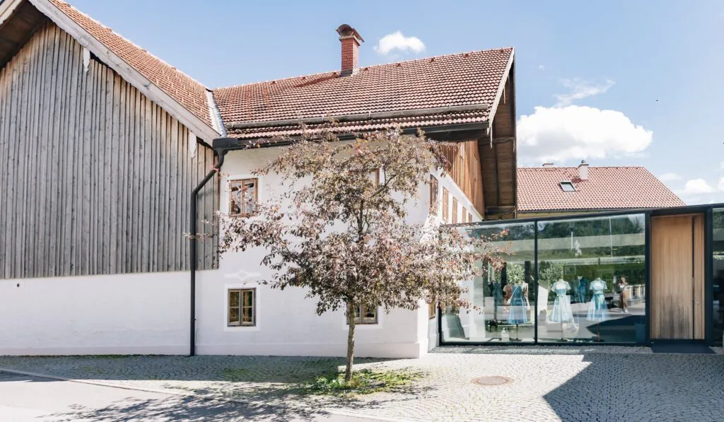 Außenansicht des Trachtenateliers Wimmer schneidert in Schleedorf – rustikales Holzgebäude mit weißer Fassade, moderner Glasanbau als Schauraum und ein blühender Baum im gepflasterten Innenhof bei Sonnenschein