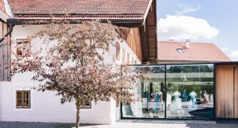 Blick auf das Trachtenatelier Wimmer schneidert in Schleedorf – ein rosa blühender Baum rahmt den modernen Glasschauraum ein, in dem Dirndl ausgestellt sind, dahinter das traditionelle weiße Bauernhaus mit Ziegeldach